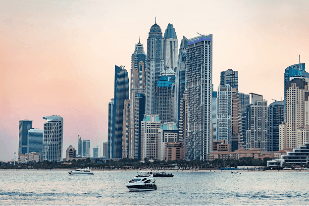 Skyline of a modern city with tall, diverse skyscrapers at dusk. Boats sail on a tranquil body of water in the foreground, under a pastel sky.